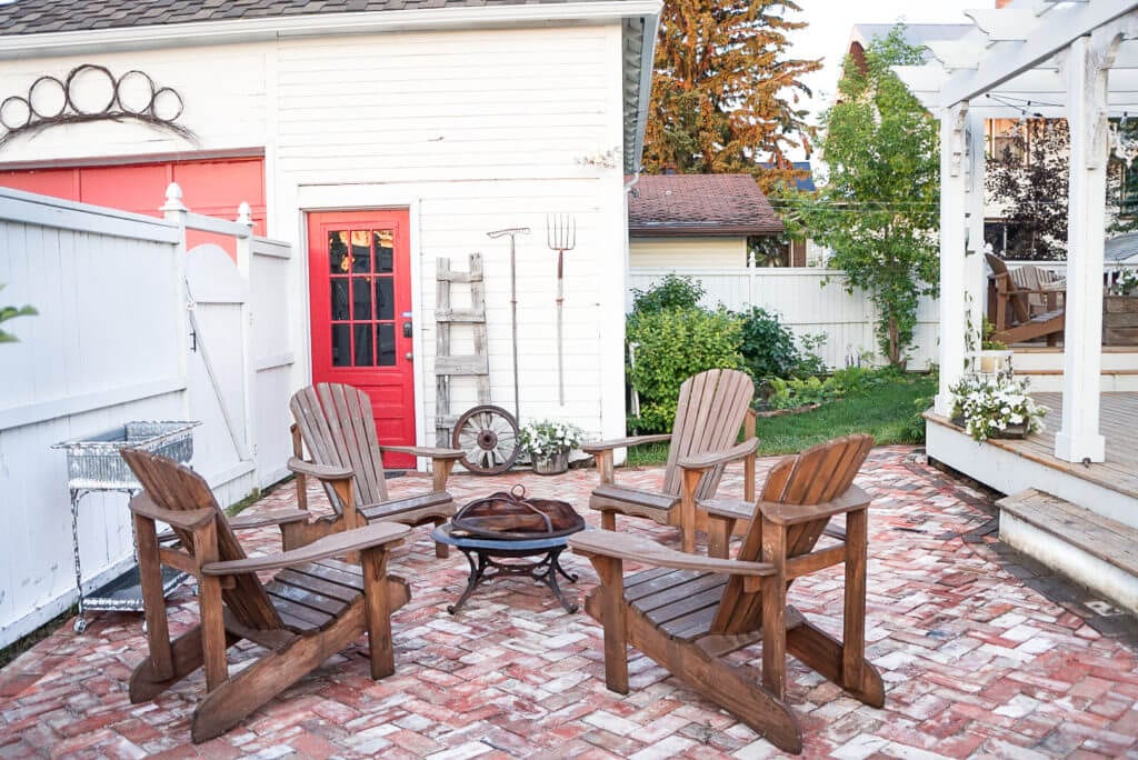 Brick patio with chairs and a fireplace beside a beautiful garden and white garage.