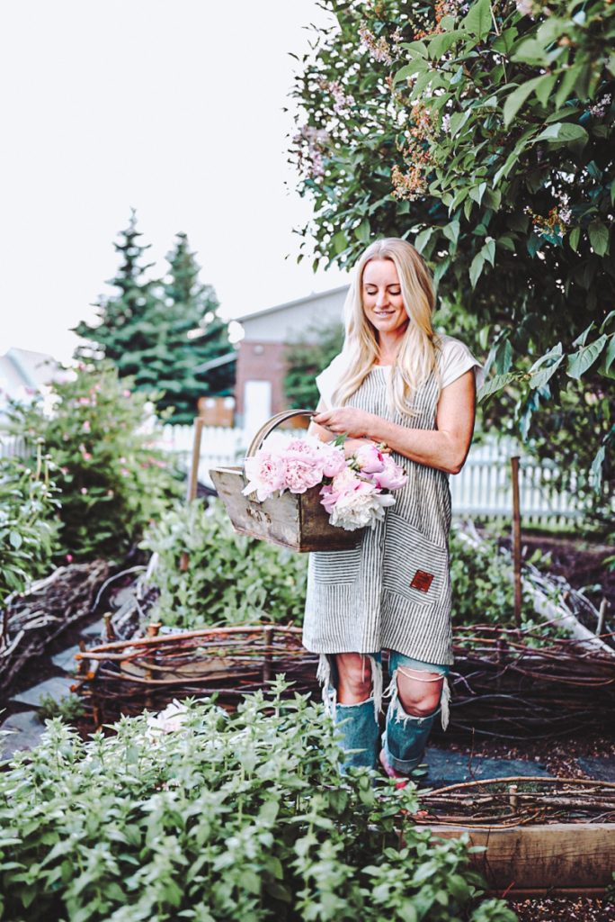 Deborah from B Vintage Style holding a wooden basket of peonies in a beautiful whimsical garden.