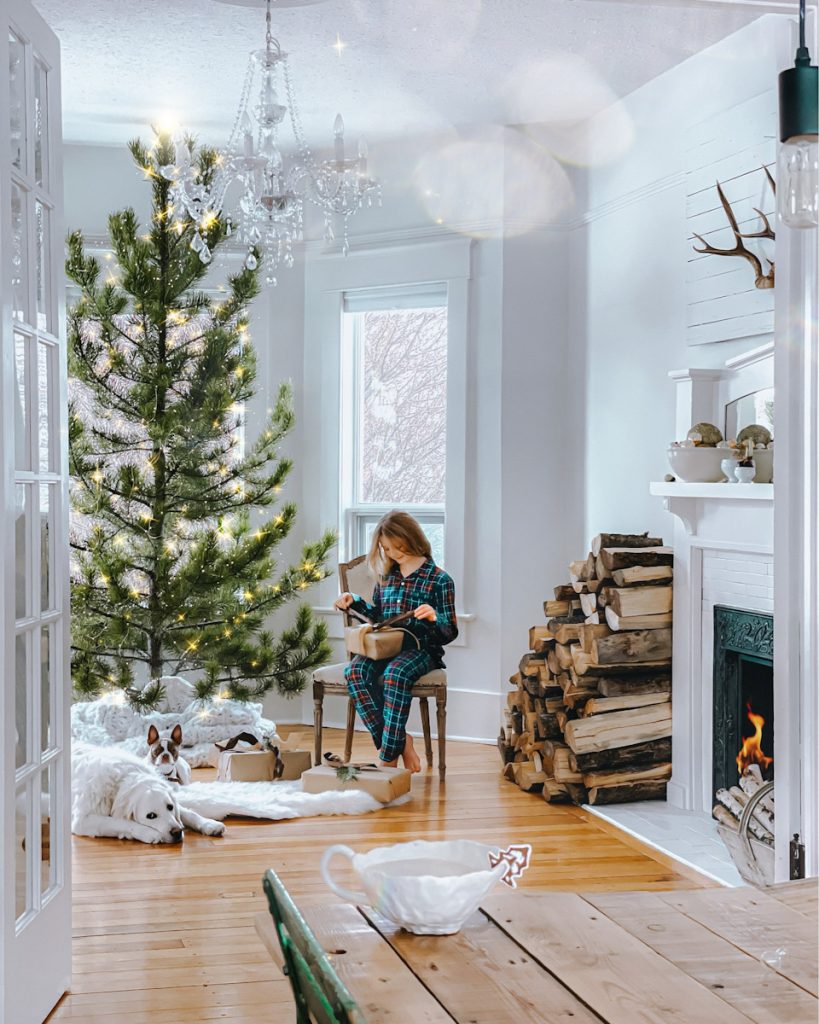 young girl wrapping a present by the fire and beside a big fresh Christmas tree that twinkles