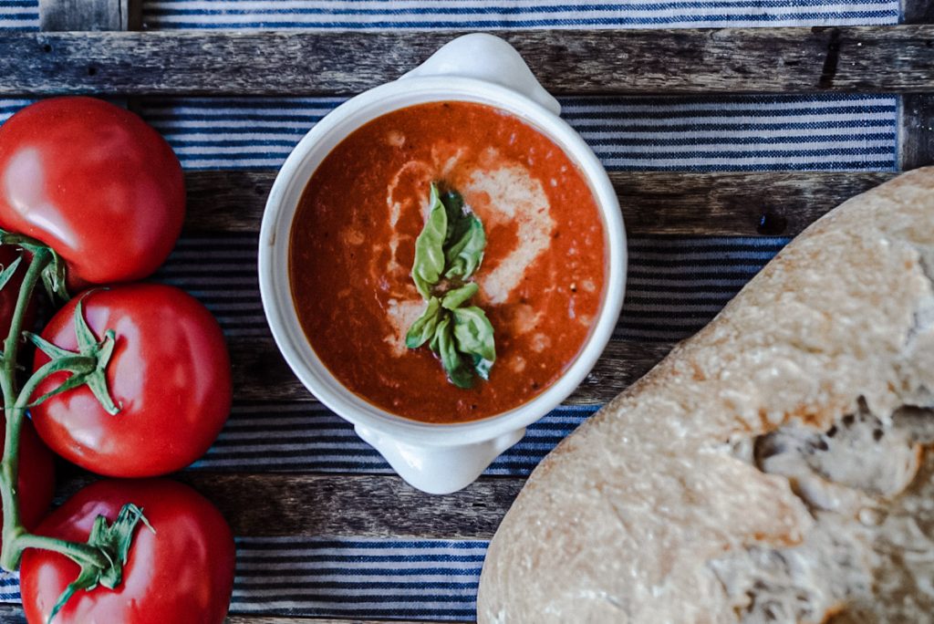Homemade tomato soup that is smoked beside fresh tomatoes and a loaf of artisan bread.