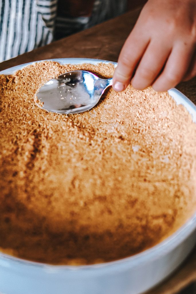 using a spoon to press a graham cracker crust into a dish to make cheesecake