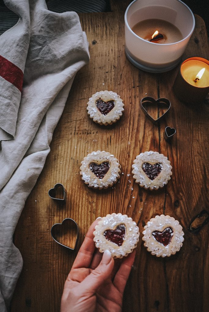 Linzer tart cookies with heart cut outs on a rustic board with heart shaped cookie cutters.