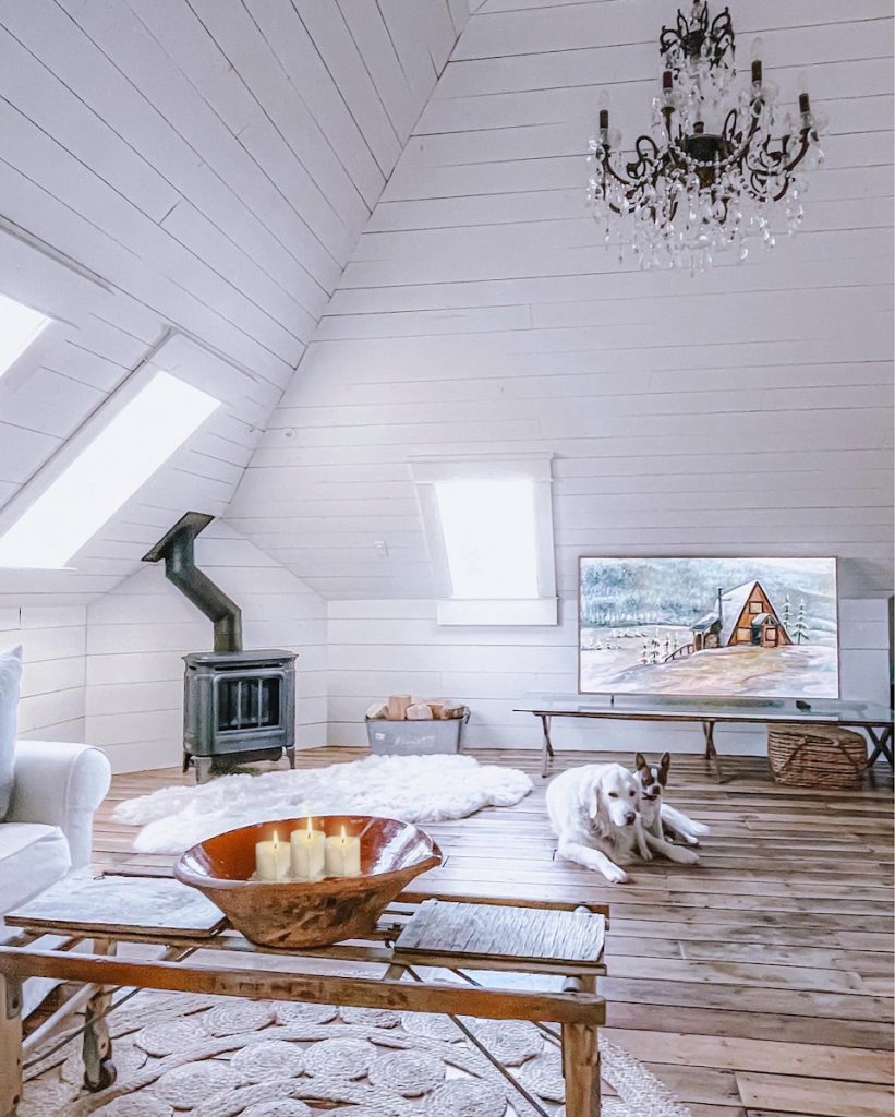 A cozy hygge styled attic space that has an antique bowl full of candles, a beautiful picture of a cozy winter cabin and a wood burning fireplace.