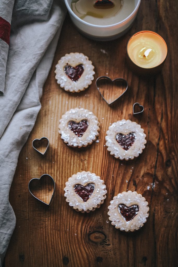 Heart cut out linzer tart cookies that are on a cutting board with heart cookie cutters and candles.
