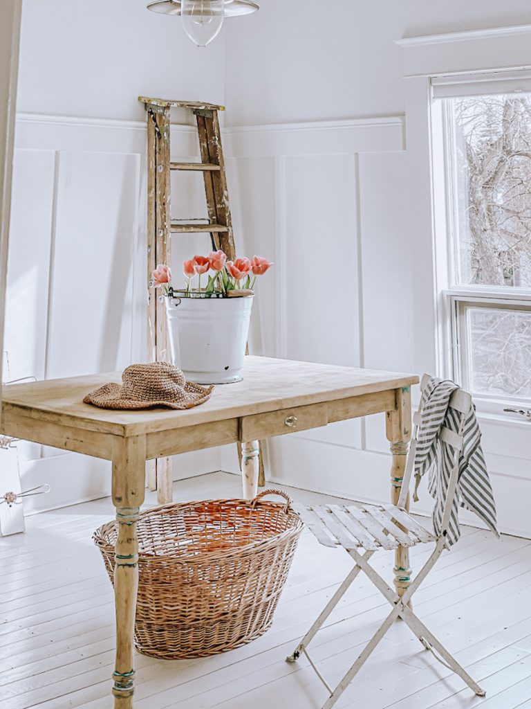 Bright office decorated in Scandinavian farmhouse decor with a raw wood desk, an antique French cafe chair, wooden ladder resting on the wall and a bucket of flowers.