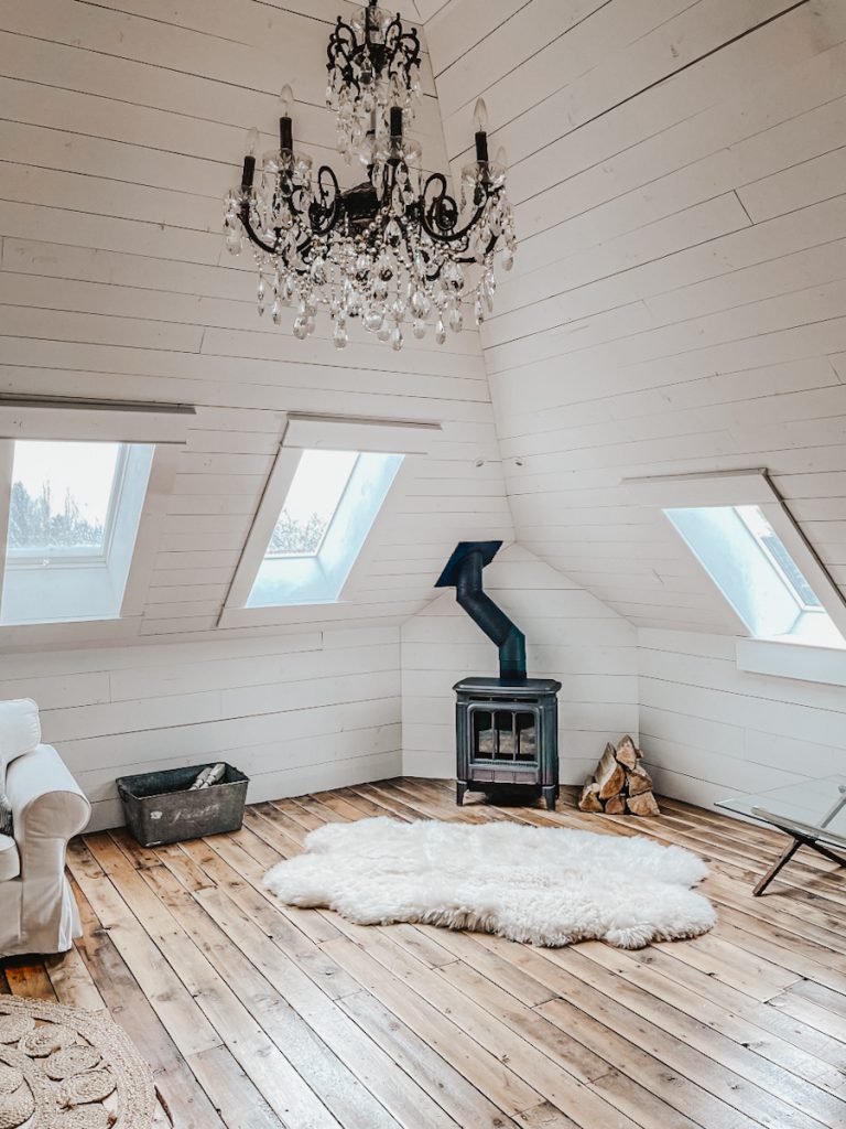 A corner of an attic family room that has a large chandelier, a black wood stove with a sheepskin rug in front of it and fire wood stacked beside it.