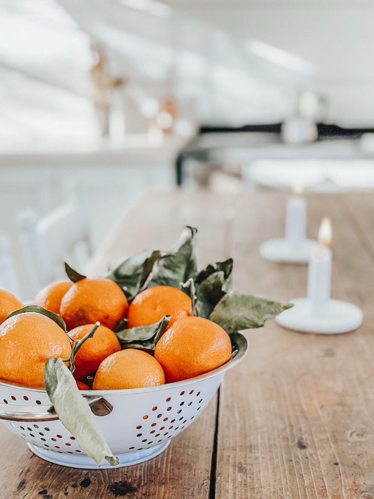 A metal strainer full of oranges with the leaved on them on a vintage table with candles.
