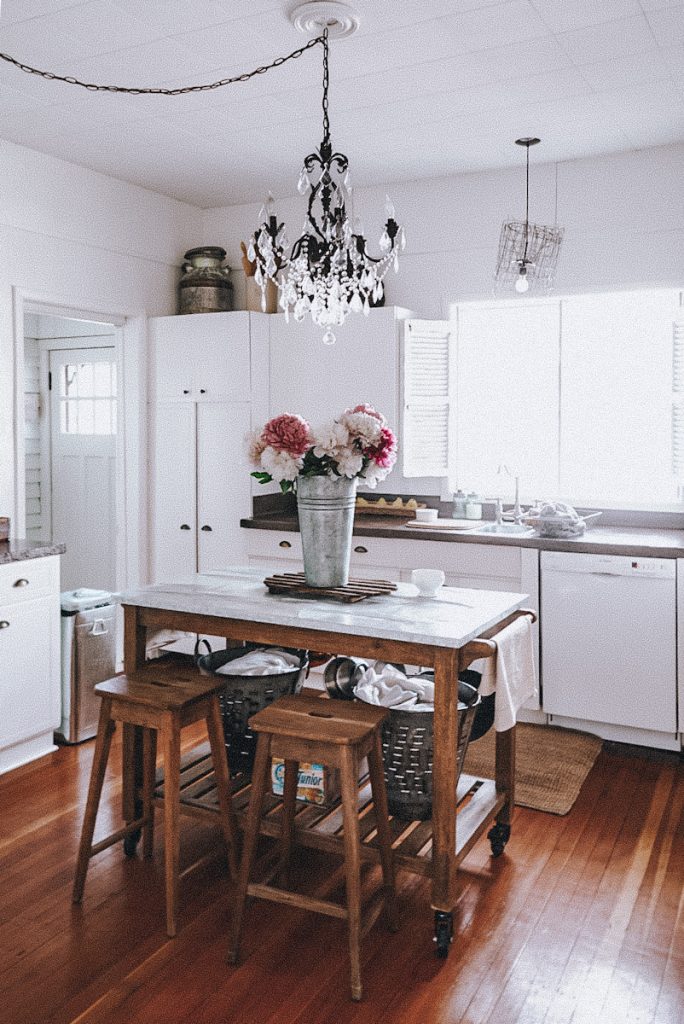 Beautiful Farmhouse kitchen in a Victorian house with a big rustic kitchen island that has a tall vase of peonies on top and a crystal chandelier above it.