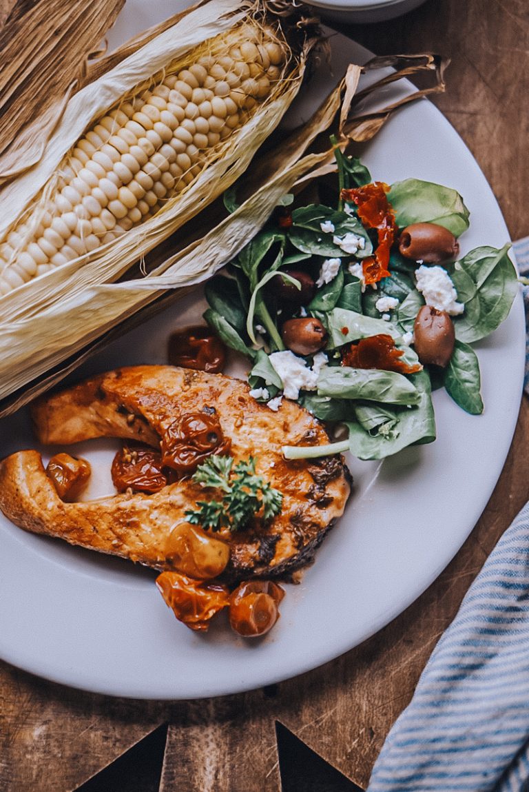 Bloody Mary Salmon on a plate with corn on the cob and a feta, olive salad.