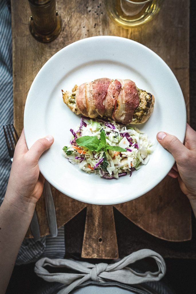Girl serving a plate with pesto stuffed chicken breast with bacon around it and a side of coleslaw.