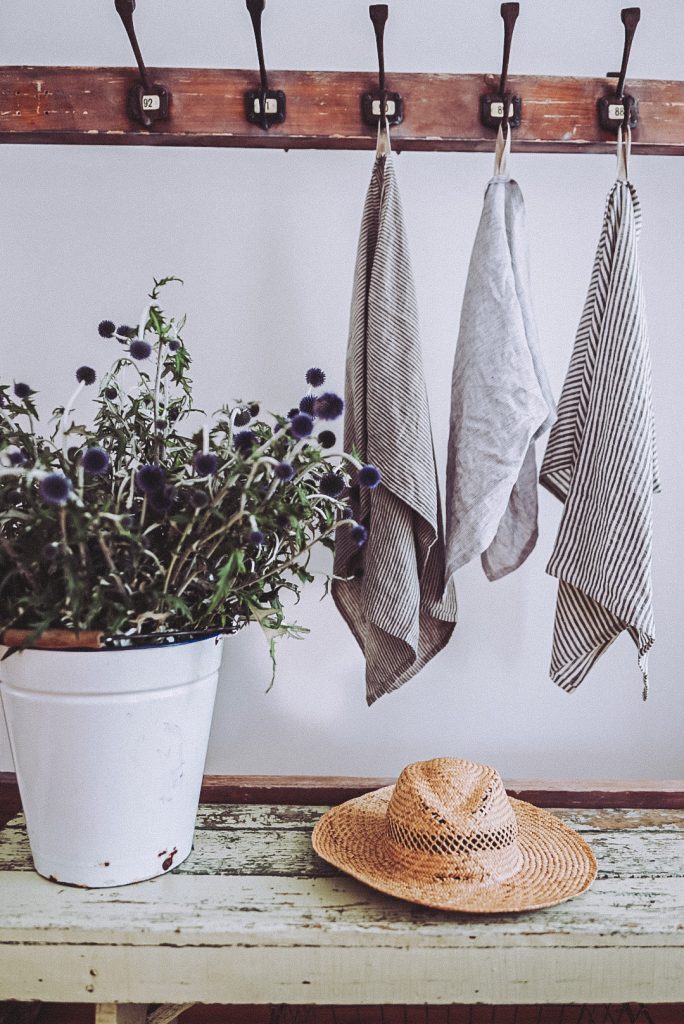 A bucket of flowers on a bench beside a hat with three linen towels hanging beside.