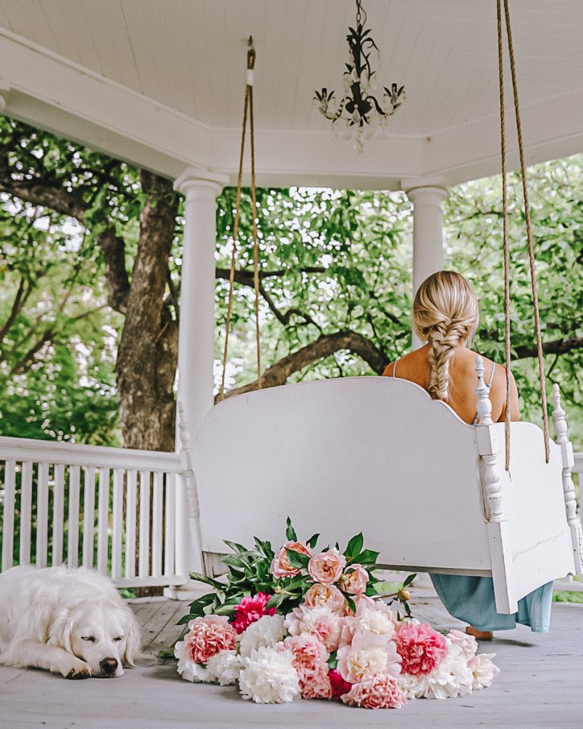 A porch swing with flowers underneath and a lady sitting on the swing.
