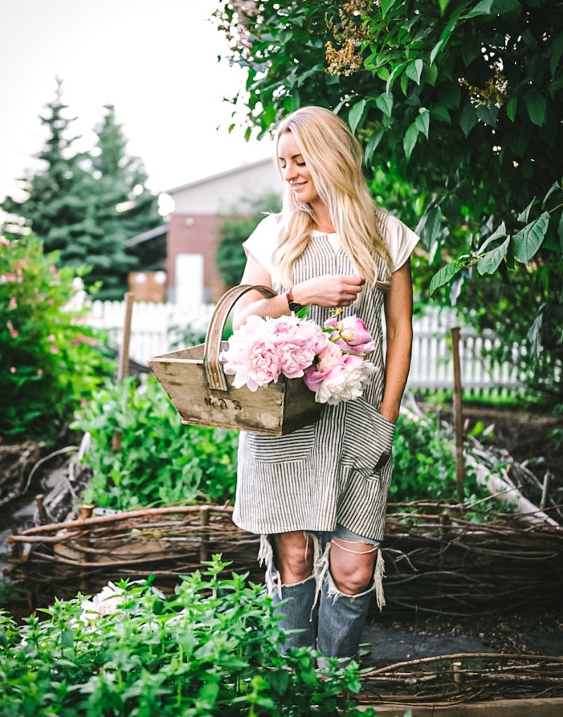 Deborah from B Vintage Style standing in a beautiful garden holding a basket of pink peonies.