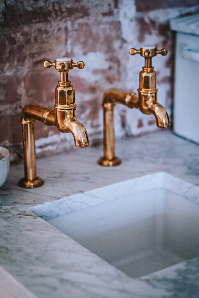 Unlacquered brass bib taps from Devol in front of a brick backsplash and marble stone countertops.