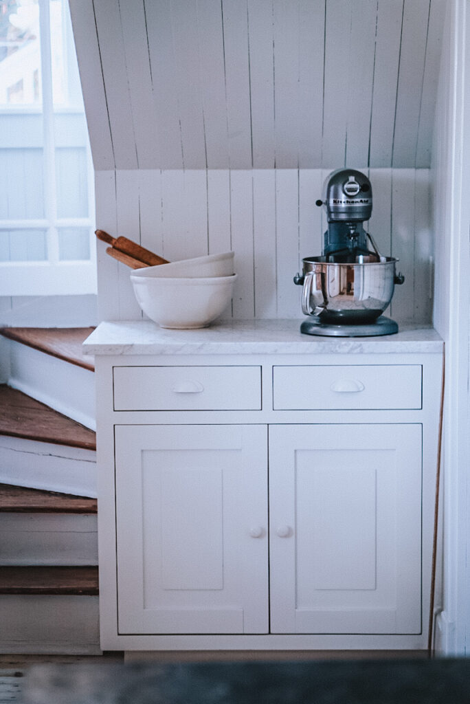 A baking station cabinet with an electic mixer, antique stoneware mixing bowls and wooden rolling pins.