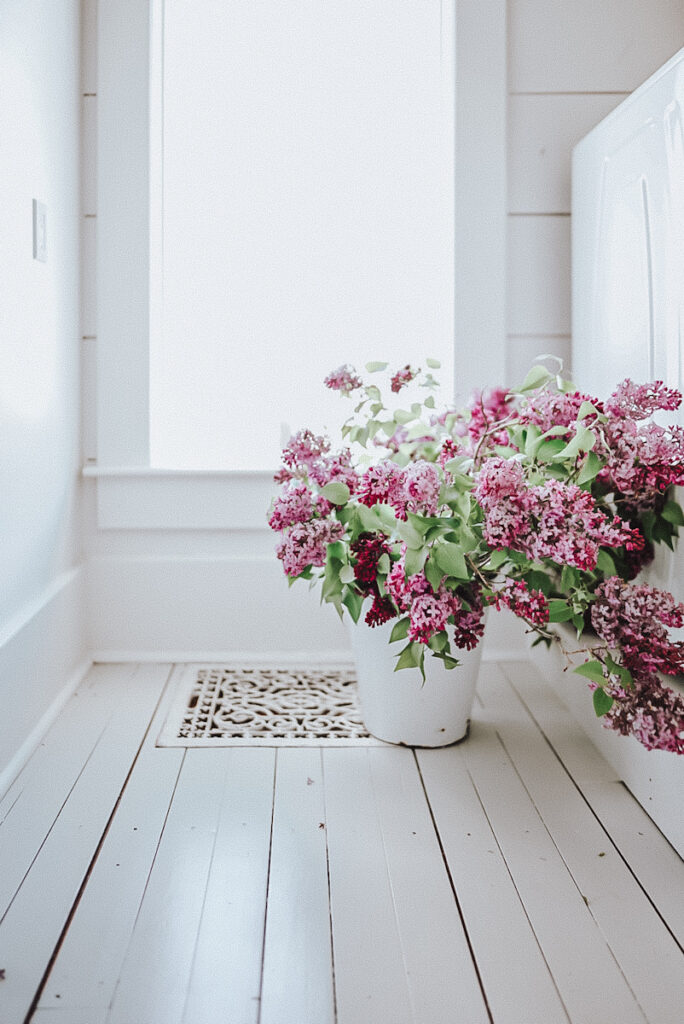 A large white enamel bucket of lilacs displayed in front of a windowsil.