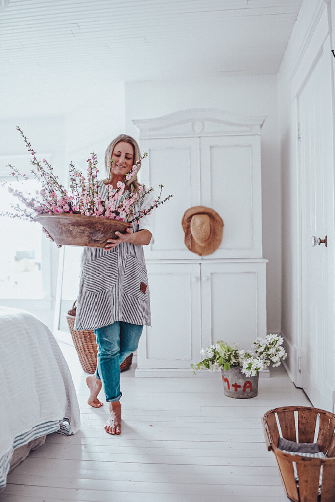 Deborah, B Vintage Style, walking with a large arrangement of blossoming flowers in a bedroom with painted floors.